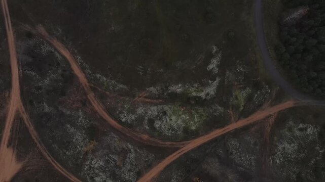 Aerial top down pan of Dretunsky training range with intersecting reddish dirt tracks, faint vehicle trails, a narrow paved road, forest edge, and a small ruined foundation.