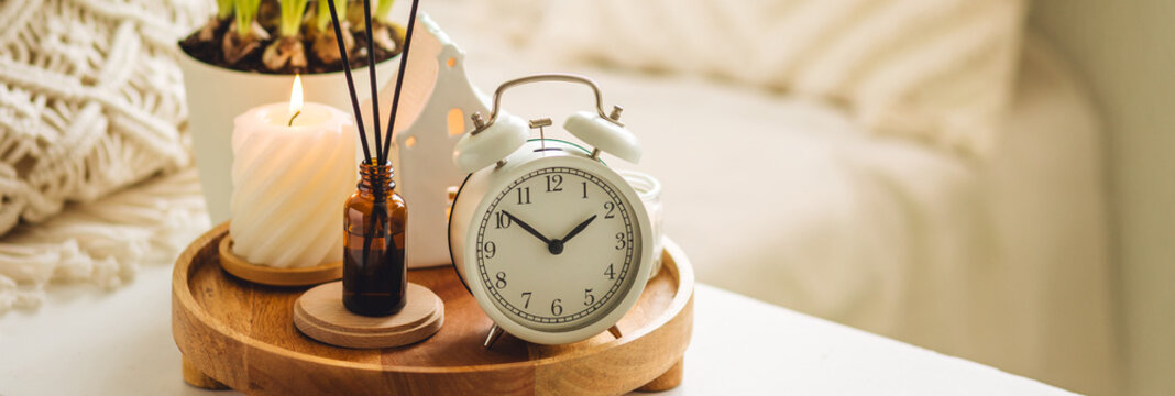 Banner. Spring change, Daylight Saving Time concept. White alarm clock, bulbous flowers, candles on the white table. Cozy home interior. Copy space