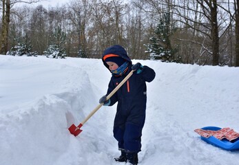 Naklejka premium Cute toddler boy dressed in warm winter clothes digging snow with a small red shovel on snowy park path. Child enjoying outdoor winter play near blue sled surrounded by deep snow and cold season.