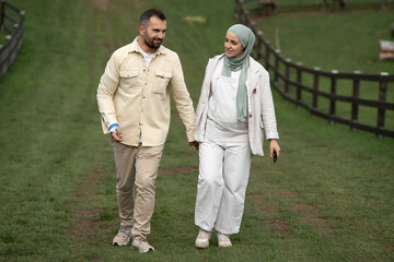 Fototapeta premium A happy couple holding hands in a lush green park, enjoying a romantic moment together, a pregnant woman in hijab, and a man with a broken arm, outdoor love story, connection, happiness, nature.