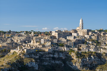 Naklejka premium Distinctive skyline of Matera, Italy, featuring clustered ancient stone buildings and a prominent cathedral tower rising above dramatic limestone cliffs under a clear blue sky.