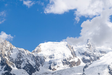 Obraz premium Sunlight bathes the pristine, snow-covered crest of Mont Blanc du Tacul, with rugged rock faces and drifting clouds set against a vivid blue alpine sky.