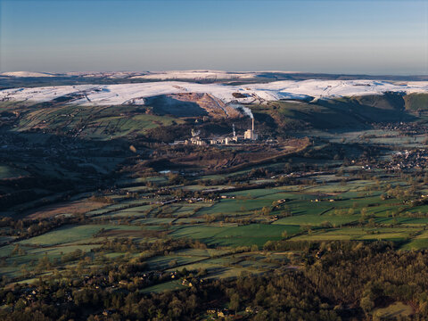 Aerial view of snow-dusted hills contrast with verdant fields, punctuated by industrial architecture, Peak District National Park, United Kingdom.