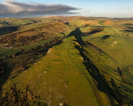 Aerial view of Mam Tor's dramatic ridge casting long shadows across the verdant valleys, Peak District National Park, United Kingdom.