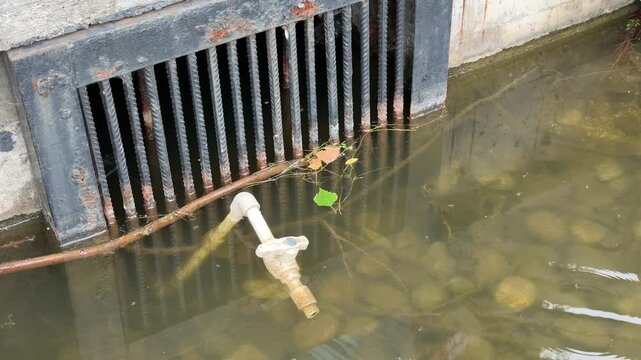 Rusty grate above pond drain, murky water swirling around exposed valve and submerged stones, small green leaf floating near grid, concrete embankment and metal pipe visible, subtle ripples.