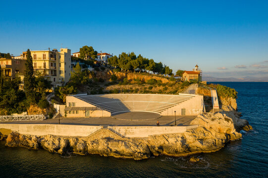 Open-air amphitheater perched on a rocky coastline in Paralio Astros, with a lighthouse and hillside houses illuminated by warm evening sunlight. The tranquil scene overlooks the deep blue sea and