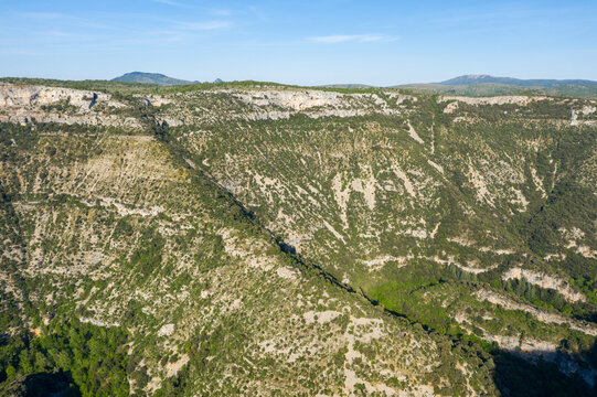 Wide view of rugged limestone canyon walls covered in scattered greenery at Cirque de Navacelles. The sunlit cliffs and deep shadows reveal dramatic natural contours beneath a clear sky.