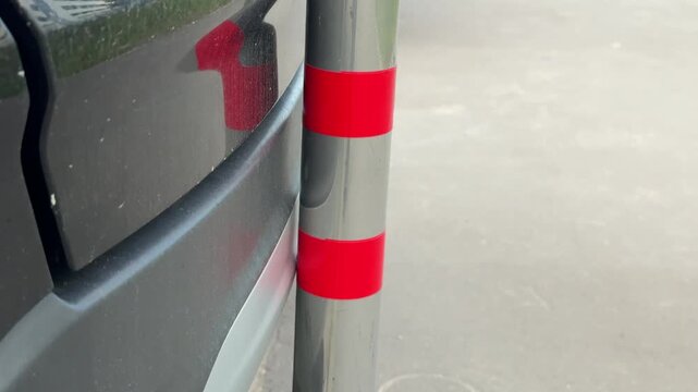 Crossover bumper scratched by metal pole, closeup rear bumper scuff on dark suv, parking edge near curb, red reflective post with paint transfer, urban outdoor light, gritty pavement.
