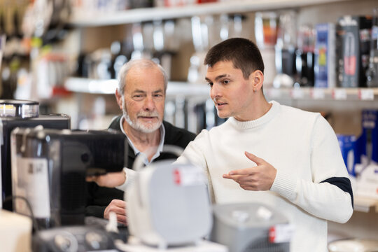Senior man with help of guy sales consultant choosing coffee machine, checks completeness of household appliances and presence of defects. Customer chooses household appliances