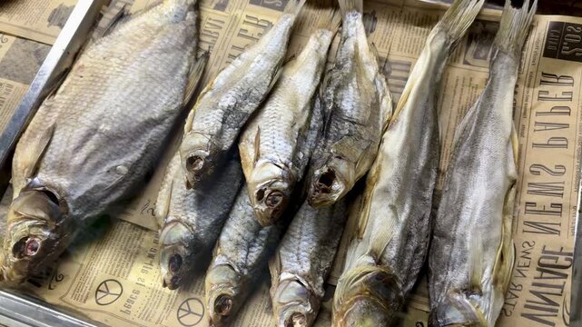 Dried fish lined on newsprint counter, salted heads arranged in tight row with golden gray scales and flaky brittle texture, closeup rustic display suggesting tavern snack sale for beer.