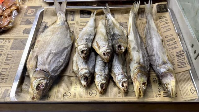 Dried fish lined on newsprint counter, salted heads arranged in tight row with golden gray scales and flaky brittle texture, closeup rustic display suggesting tavern snack sale for beer.