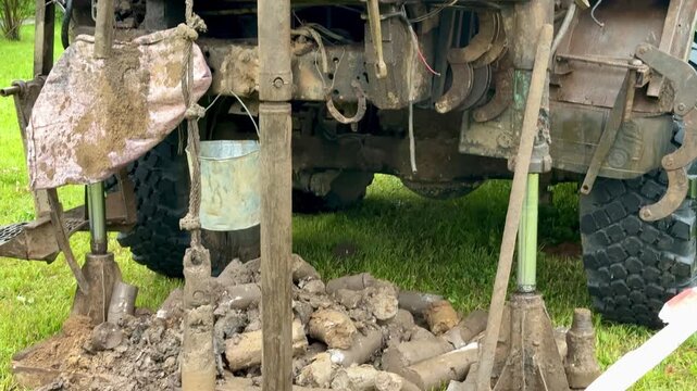 Tracked drilling rig extracting soil cores on grassy field, metal tracks visible, auger drilling into ground, columns of extracted soil stacked nearby, muddy site conditions under overcast.