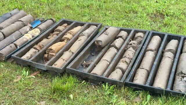 Soil core samples laid on grass drilling rig and auger near shallow borehole muddy cylindrical cores in plastic trays shovel and drill bits beside borehole geologist performing field logging.