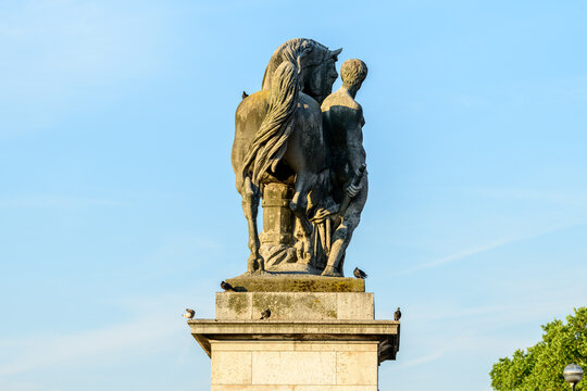 Rear view of a classical stone sculpture depicting a man and a horse atop a pedestal on Pont de Iena in Paris, illuminated by warm sunlight against a clear blue sky.