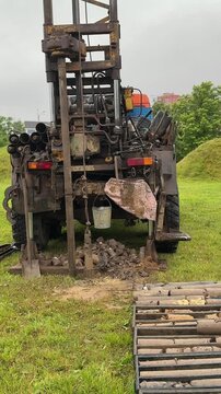 Tracked drilling rig extracting soil cores on grassy field, metal tracks visible, auger drilling into ground, columns of extracted soil stacked nearby, muddy site conditions under overcast.