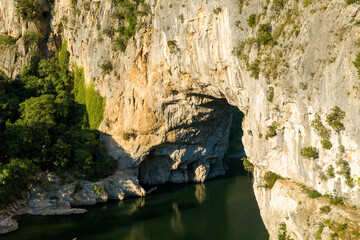 Golden sunlight illuminates the massive limestone arch of Pont d'Arc as it towers above a calm river, with lush green trees and rugged cliffs creating dramatic contrasts in the Ardeche region.