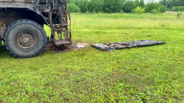 Drilling rig extracting soil cores in grassy field, truck mounted rotary rig creating borehole while driller operates controls, core tray with cylindrical samples laid on grass, geologist.