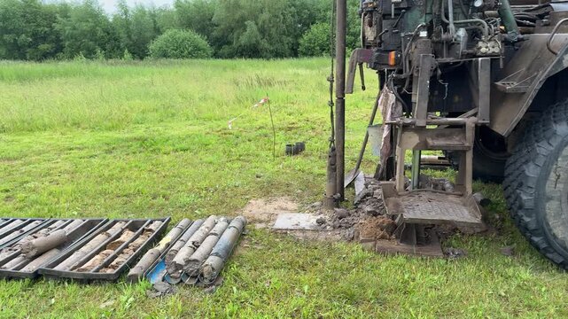Drilling rig extracting soil cores in grassy field, truck mounted rotary rig creating borehole while driller operates controls, core tray with cylindrical samples laid on grass, geologist.
