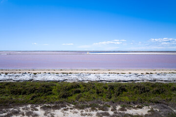 Striking view of pink saltwater ponds bordered by white mineral crust and green marsh vegetation beneath a vast, cloud-streaked blue sky in Camargue. The scene is open and vibrant, highlighting unique