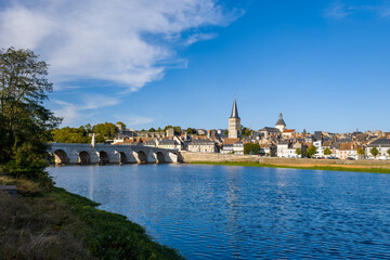 Naklejka premium Clear morning sunlight illuminates the historic stone bridge and traditional rooftops of La Charite sur Loire, with the church spire rising above the riverside town. Calm blue water and lush greenery