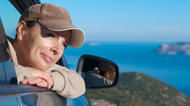 Happy woman enjoying a scenic road trip. Serene woman in a baseball cap leaning out of a car window, smiling while admiring the breathtaking coastal scenery during a sunny day