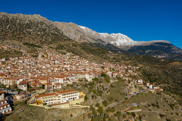 Obraz premium Aerial view of Arachova, Greece, showing a dense cluster of red-roofed houses spread across a rugged hillside, with dramatic snow-capped mountains rising in the background under a vivid blue sky.