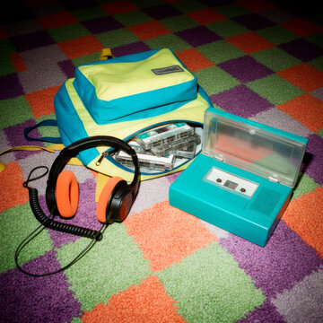 90s style photo: a open backpack lies on the floor, cassettes, headphones with foam ear cushions and a portable player spill out of it, the texture of a carpet with a geometric pattern, camera flash