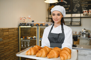 Baker woman holding tray of freshly baked croissants in bakery shop