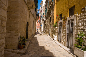 Obraz premium Narrow cobblestone street lined with textured limestone buildings and colorful facades in the historic center of Trani, Italy. Potted plants and balconies add charm to the quiet Mediterranean scene