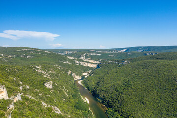 Expansive aerial view of the Gorges de l'Ardeche, featuring a winding river cutting through densely forested hills under a clear blue sky. Sunlight enhances the vibrant greenery and rugged limestone