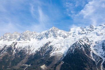 Jagged summit of Aiguille du Midi rises above the sweeping Glacier des Bossons, with snow-covered slopes and scattered forests beneath a dynamic blue sky.