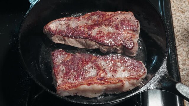 Two New York strip steaks searing in a hot cast iron skillet