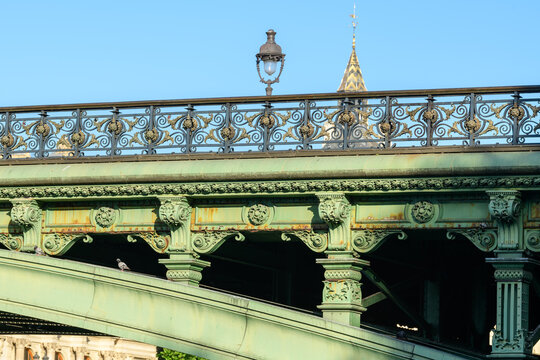 Decorative green ironwork and intricate railing of Pont Notre-Dame in Paris, highlighted by afternoon sunlight with a clear blue sky and architectural spire in the background.