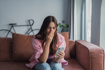 Obraz premium A frustrated millennial Latin woman sits on the sofa reading sudden bad news on her phone, covering her face with her hand in despair after receiving a threatening message or upsetting email