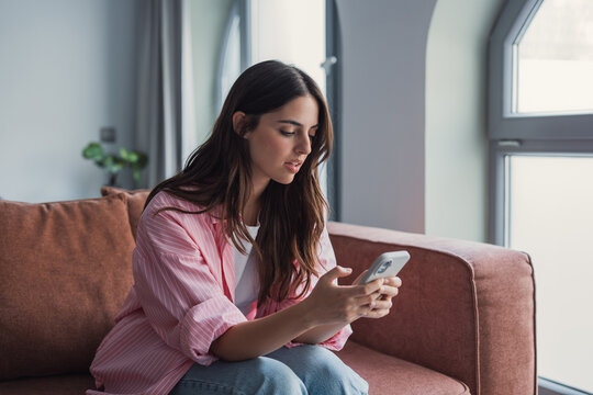 A confused and angry young woman sits on the couch at home looking at her smartphone, feeling frustrated by a broken device or bad news in a message that disrupts her daily routine