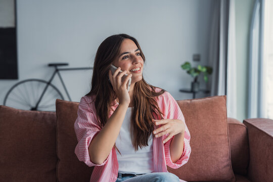 A happy woman relaxes on the couch while talking on her smartphone, smiling as she shares news with family and enjoys a pleasant personal conversation using modern mobile communication at home