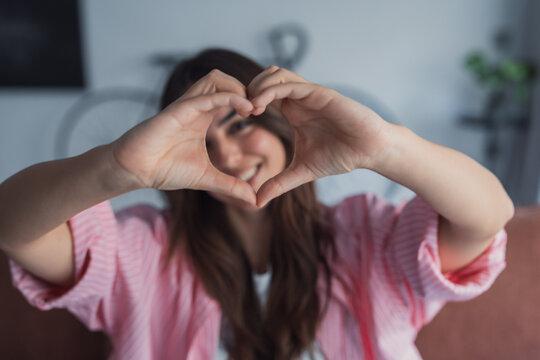 An attractive optimistic woman forms a heart sign with her hands while looking at the camera, showing affection, warmth, and emotional connection in a bright lifestyle portrait