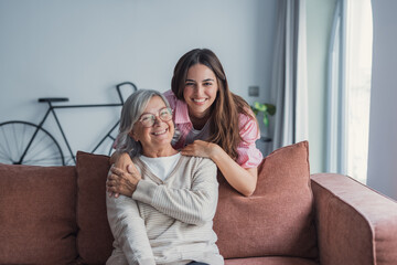 An adult daughter stands behind the couch at home hugging her older mother, sharing a tender family moment while looking at each other with affection and warmth across two generations © Daniel