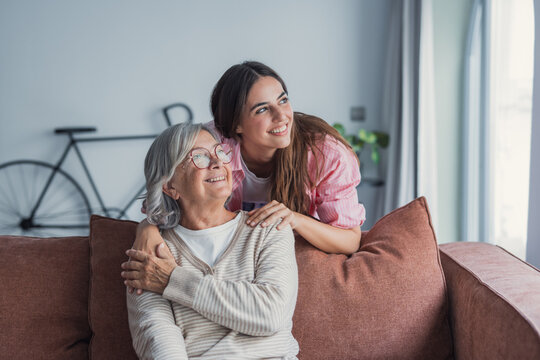 Loving adult daughter hugging older mother, standing behind couch at home, family enjoying tender moment together, young woman and mature mum or grandmother looking at each other, two generations