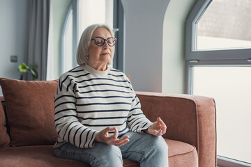 Obraz premium A mindful mature woman practices meditation at home, sitting calmly on a couch in a relaxed pose while breathing deeply and enjoying a stress-free weekend focused on mental balance and wellbeing