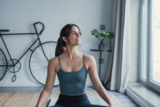 A brunette woman sits on the floor at home on a carpet wearing sportswear, practicing yoga while looking outside with a gentle smile, enjoying a peaceful moment of relaxation and mindful movement