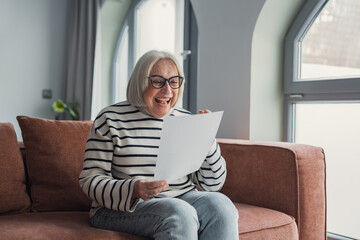 Obraz premium Happy elderly 60s excited about good news, reading paper mail letter in living room. Senior woman receiving bank notice about loan or savings, income from investment, getting post correspondence&nbsp;