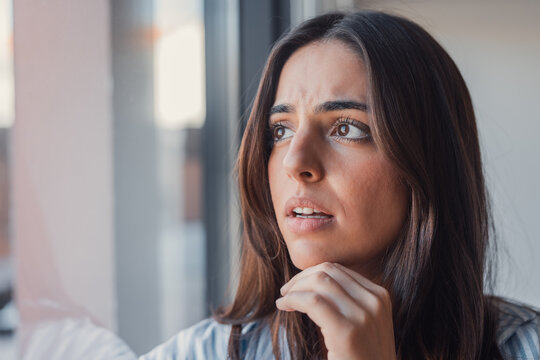 A thoughtful young woman gazes out the window with a concerned expression, captured in a casual portrait that represents uncertainty, decision making, and emotional reflection at home