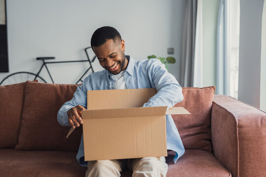 Smiling African American cheerful man unpacking parcel, sitting on couch at home, happy satisfied customer looking into cardboard box, received online store order, good delivery service
