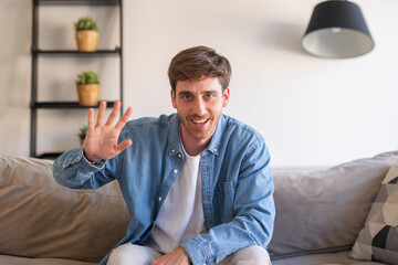 Fototapeta premium A young man smiles at the camera during an online call from his living room sofa, representing modern communication, social connection, and a friendly digital lifestyle