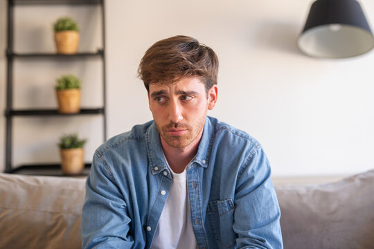 An upset millennial man sits alone on a sofa, reflecting on personal problems and grief, showing vulnerability, sadness, and the weight of negative emotions in a silent home environment