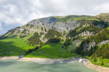 Fototapeta premium Steep green hills dotted with coniferous trees rise above a calm mountain reservoir, with rocky outcrops and scattered clouds adding depth to the scenic landscape near Barrage de Saint Guerin.