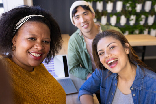 Diverse coworkers smiling for group selfie around wooden table with laptop chart in modern office