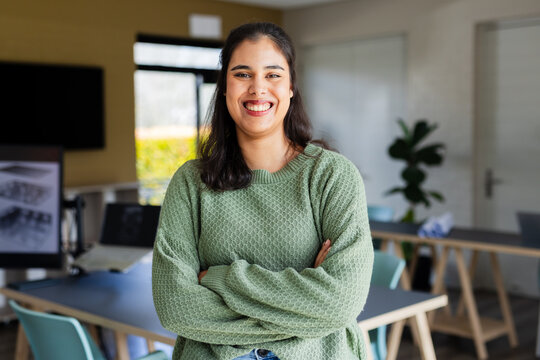 South Asian woman standing with crossed arms smiling in office meeting room with laptop