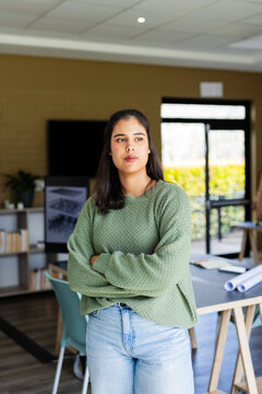 Woman standing with arms crossed wearing green sweater and jeans in design studio near blueprints